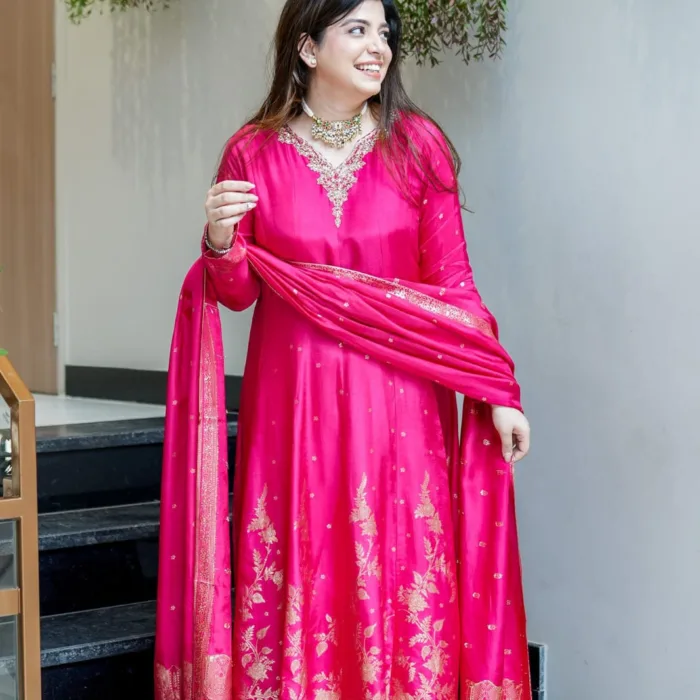 A woman in a vibrant rani pink Anarkali suit featuring gold Banarasi zari weaving at the flare and an intricate hand-worked neckline, paired with a matching pink dupatta.