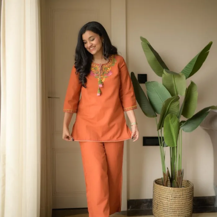 A woman posing in a Rust Orange Cotton Slub Premium embroidered co-ord set in rust orange cotton slub co-ord set featuring colorful floral embroidery on the neckline with hanging tassels and matching straight pants.
