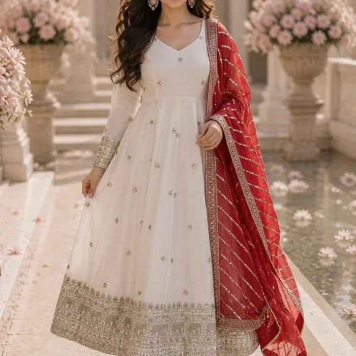 A woman posing in a featuring a white Vichitra White Anarkali Gown with gold embroidery at the hem and a contrasting red georgette dupatta with sequence work.